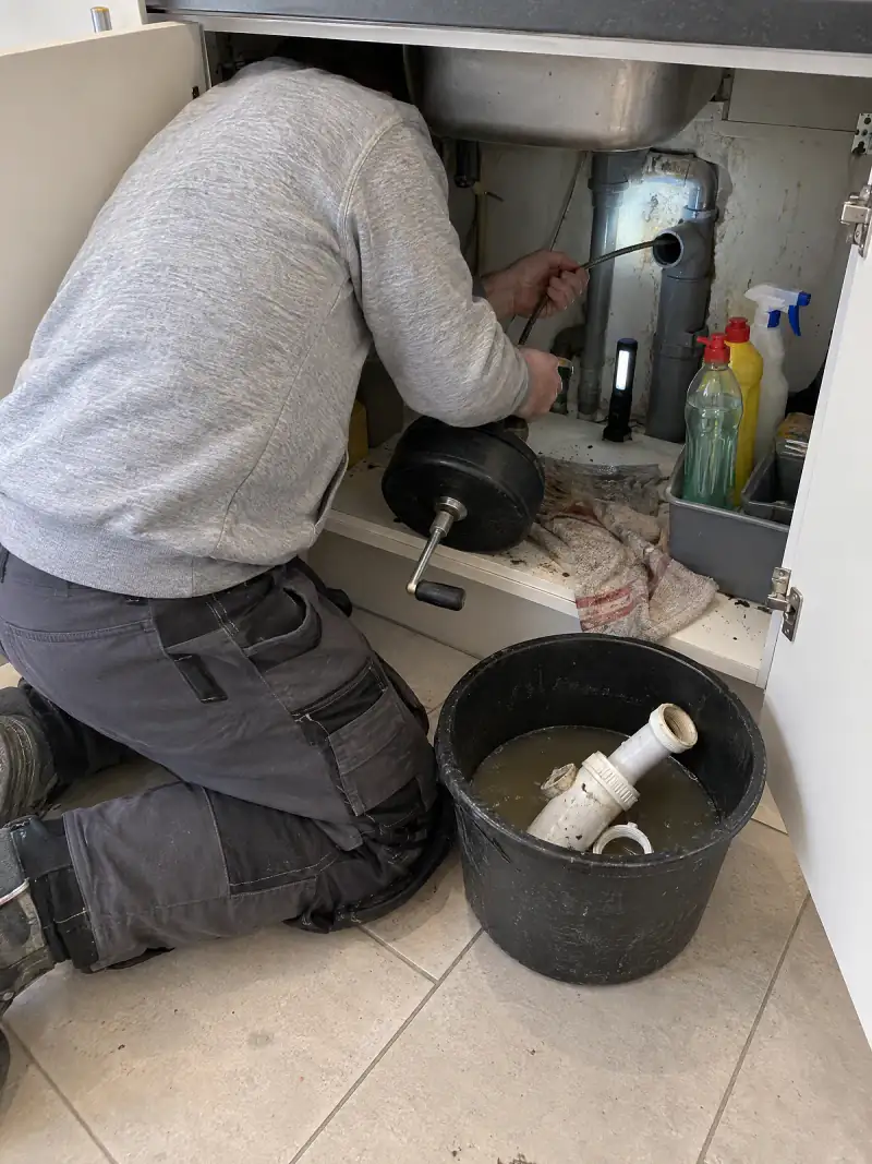 Sous un évier : un technicien passe le câble d'un furet dans l'évacuation, le siphon démonté dans un seau d'eau sale